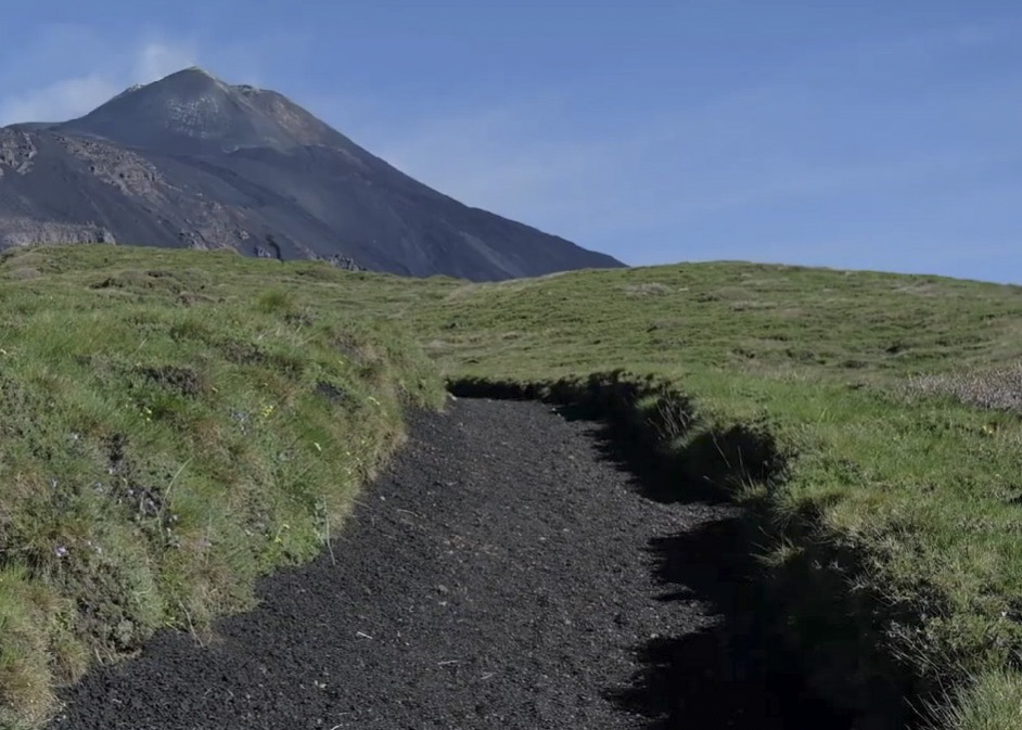 Sentiero di terreno vulcanico tra la vegetazione con l’Etna sullo sfondo